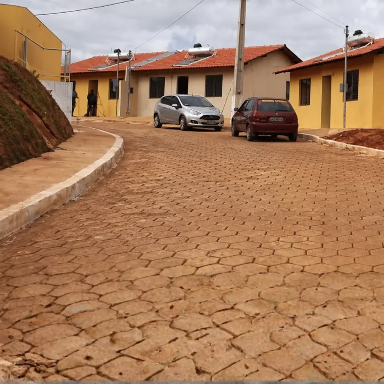 Rua pavimentada com blocos intertravados em conjunto habitacional com casas térreas amarelas e carros estacionados.