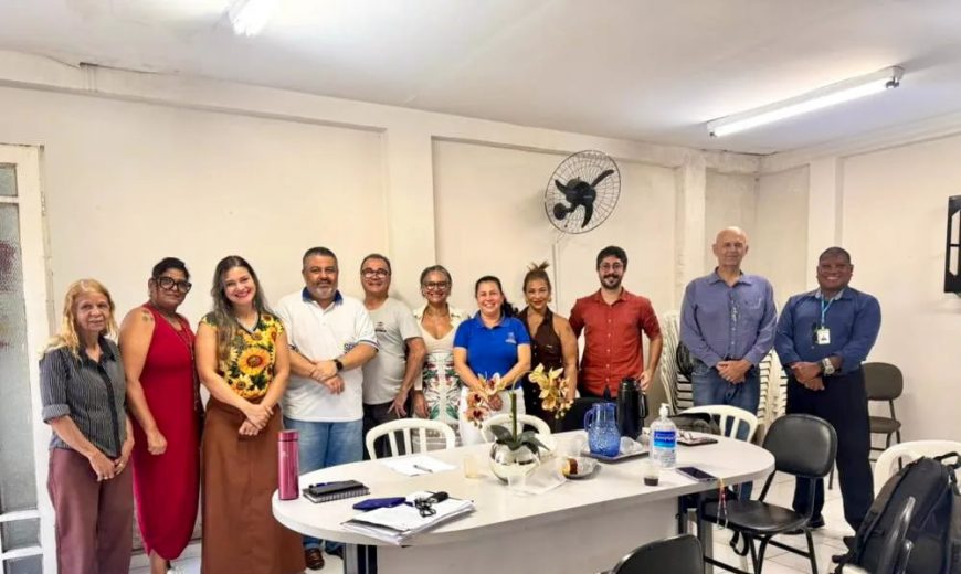 Integrantes do Conselho de Habitação posam em grupo durante reunião em uma sala simples, com mesa ao centro, cadeiras e materiais de trabalho
