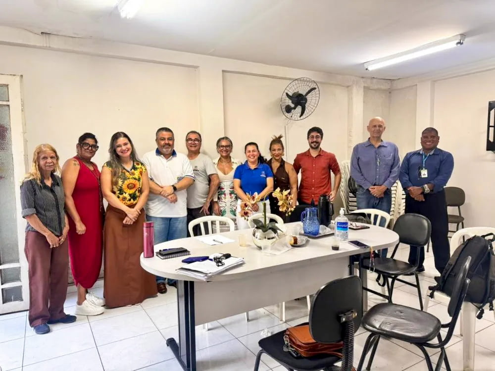 Integrantes do Conselho de Habitação posam em grupo durante reunião em uma sala simples, com mesa ao centro, cadeiras e materiais de trabalho