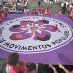 Mulheres seguram grande bandeira roxa do Coletivo de Mulheres da Central de Movimentos Populares durante atividade coletiva em espaço amplo.