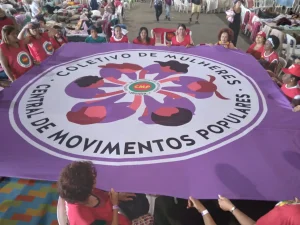 Mulheres seguram grande bandeira roxa do Coletivo de Mulheres da Central de Movimentos Populares durante atividade coletiva em espaço amplo.