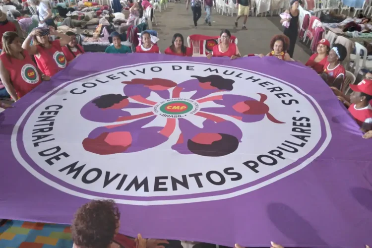 Mulheres seguram grande bandeira roxa do Coletivo de Mulheres da Central de Movimentos Populares durante atividade coletiva em espaço amplo.