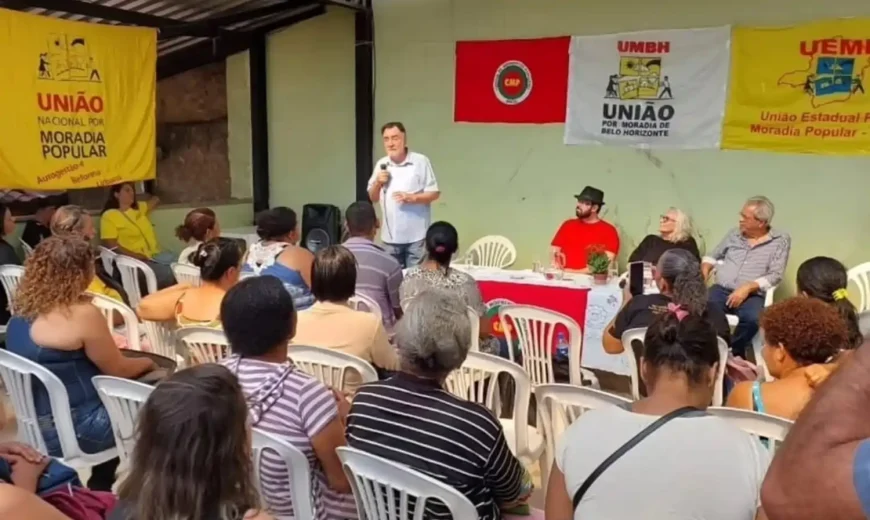 Participantes acompanham reunião da União por Moradia Popular em Belo Horizonte, com fala de um representante ao microfone e faixas de movimentos de moradia ao fundo.
