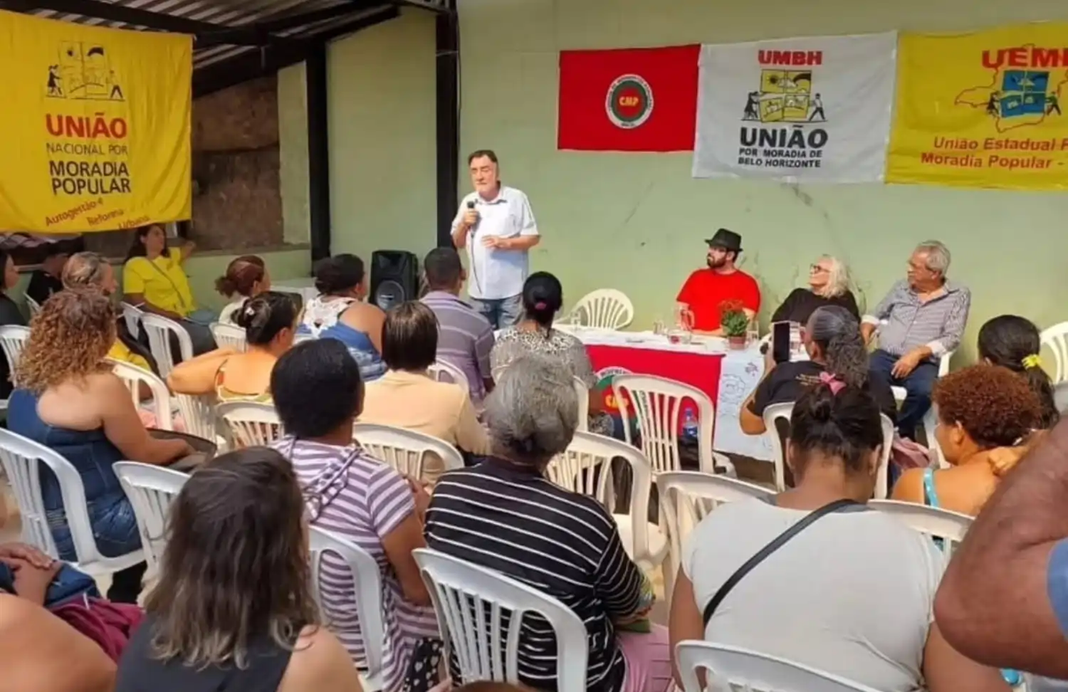 Participantes acompanham reunião da União por Moradia Popular em Belo Horizonte, com fala de um representante ao microfone e faixas de movimentos de moradia ao fundo.