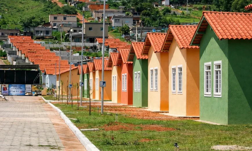 Fileira de casas populares coloridas com telhados cerâmicos em conjunto habitacional, ao lado de via pavimentada e área gramada.