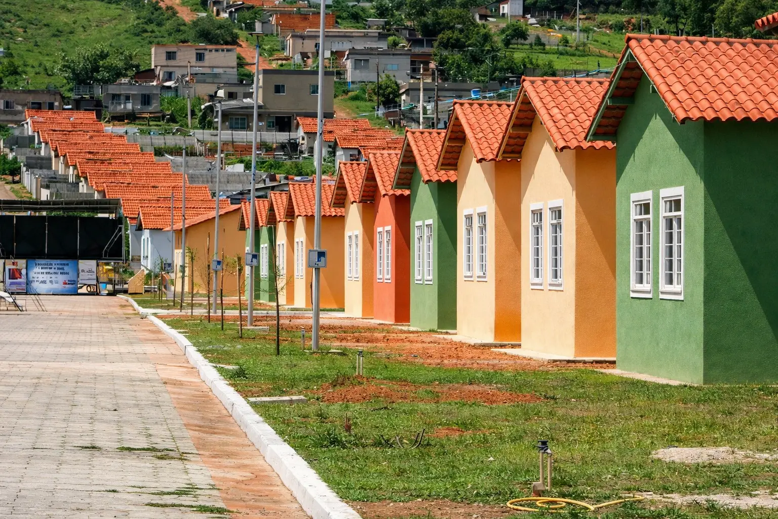 Fileira de casas populares coloridas com telhados cerâmicos em conjunto habitacional, ao lado de via pavimentada e área gramada.