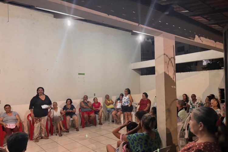 Moradores participam de reunião comunitária sobre moradia popular em espaço coberto, com cadeiras em círculo e fala de uma participante ao centro