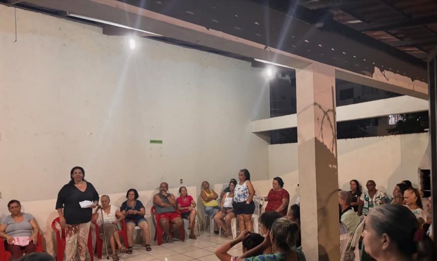 Moradores participam de reunião comunitária sobre moradia popular em espaço coberto, com cadeiras em círculo e fala de uma participante ao centro