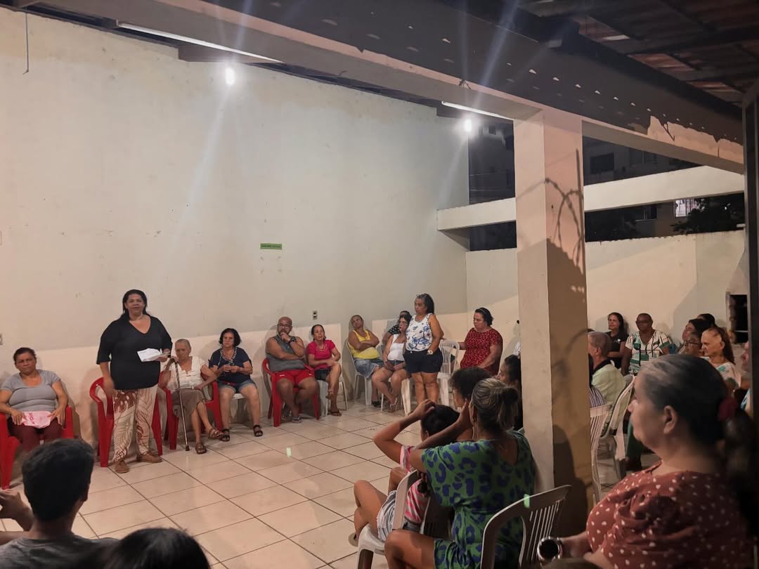 Moradores participam de reunião comunitária sobre moradia popular em espaço coberto, com cadeiras em círculo e fala de uma participante ao centro